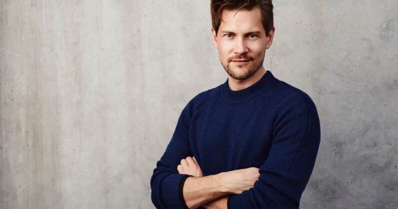 Mann im dunkelblauen Pullover steht mit verschränkten Armen vor einer grauen Wand.