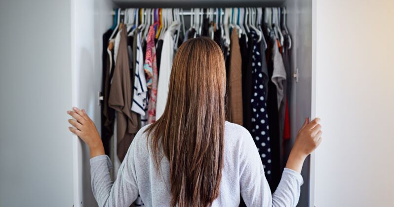 Frau von hinten blickt in einen offenen Kleiderschrank mit vielen Kleidungsstücken auf Bügeln.