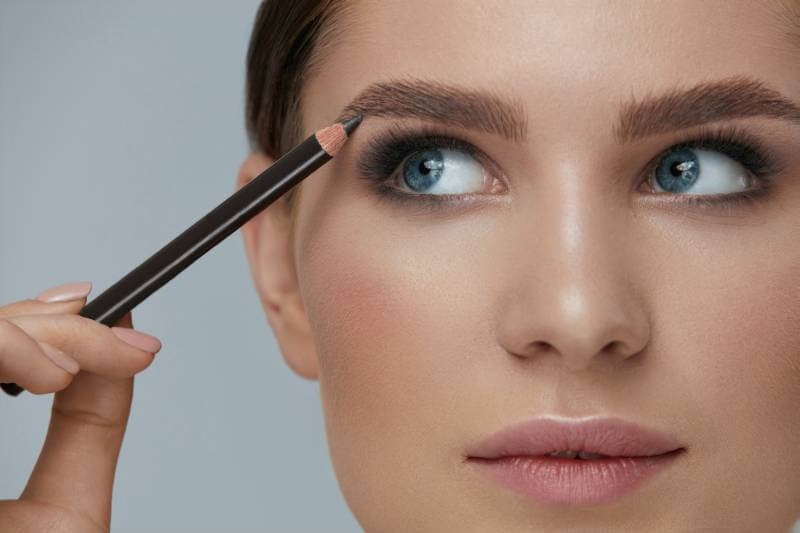 Nahaufnahme eines Gesichts: Make-up wird mit einem Stift an der Augenbraue aufgetragen.